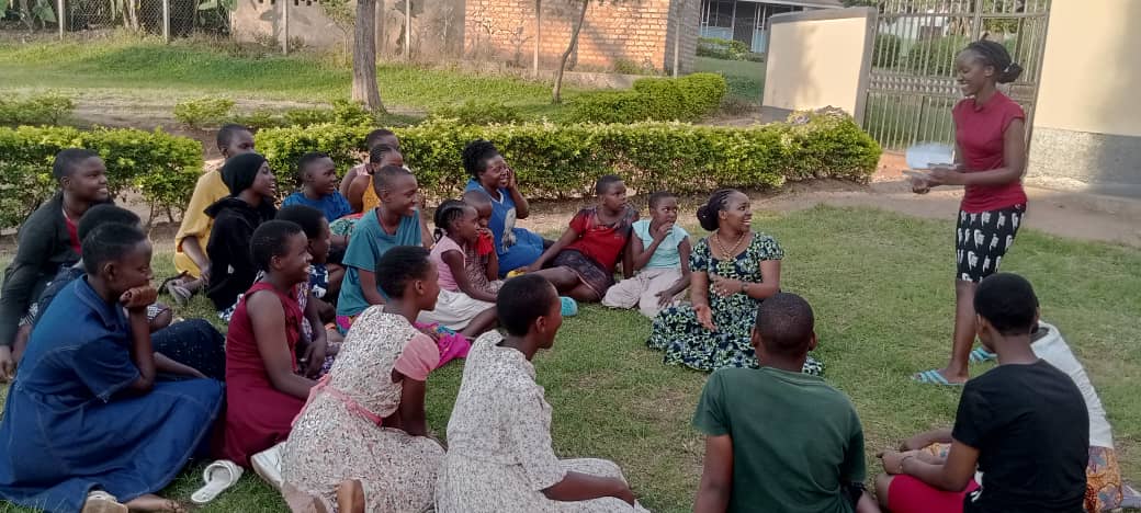 Teen girls seated on the ground, looking with smiles at a young woman who is speaking