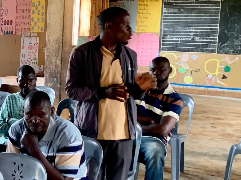 Man standing in profile talking to other seated men in a class